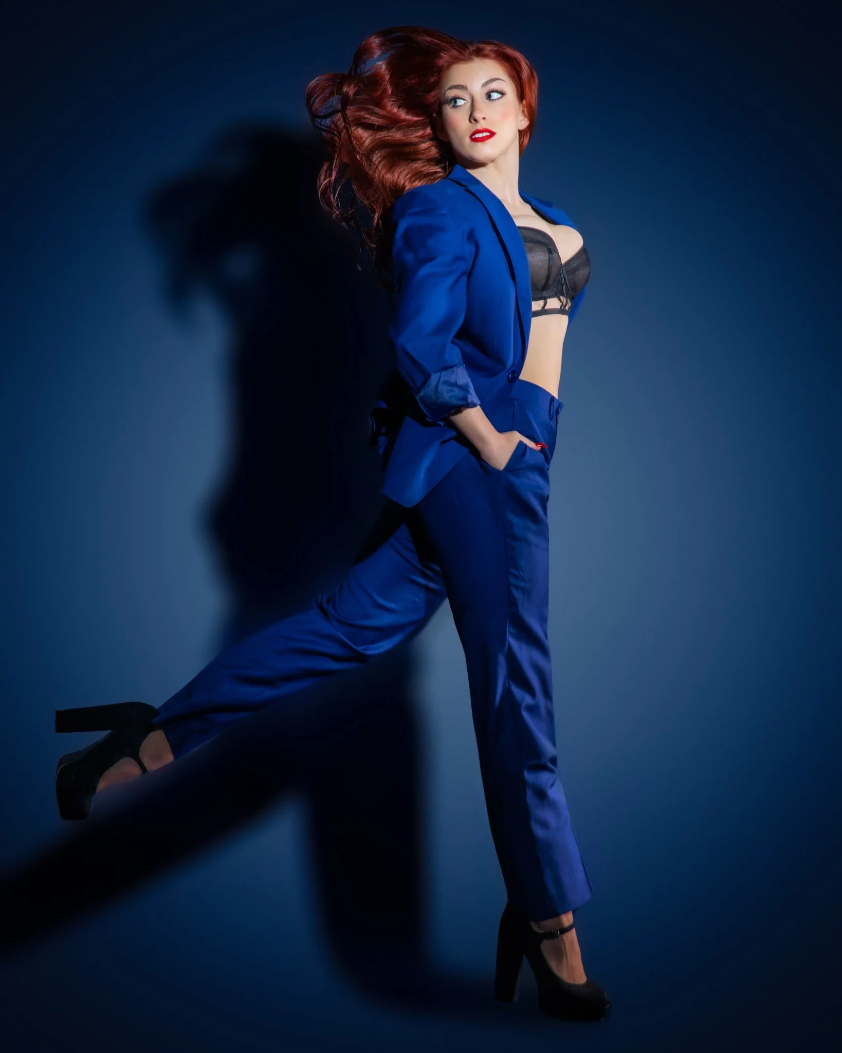 A woman with long red hair in a pose against a dark background, wearing a blue suit.